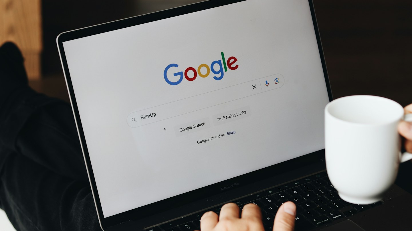 Person using Google Search on a laptop while holding a white coffee mug.