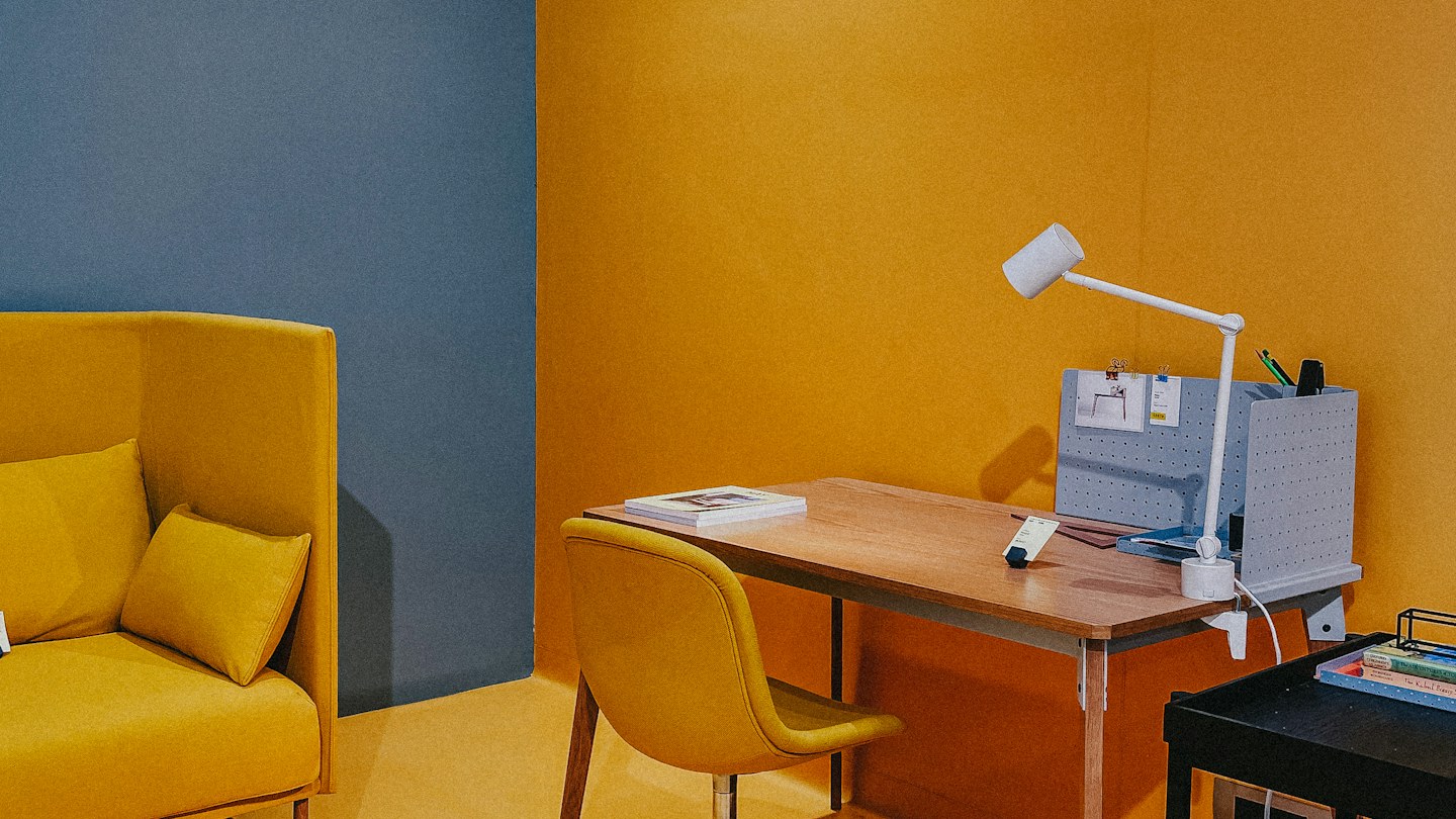 Creative workspace with a yellow chair, mustard wall, wooden desk, and white task lamp.